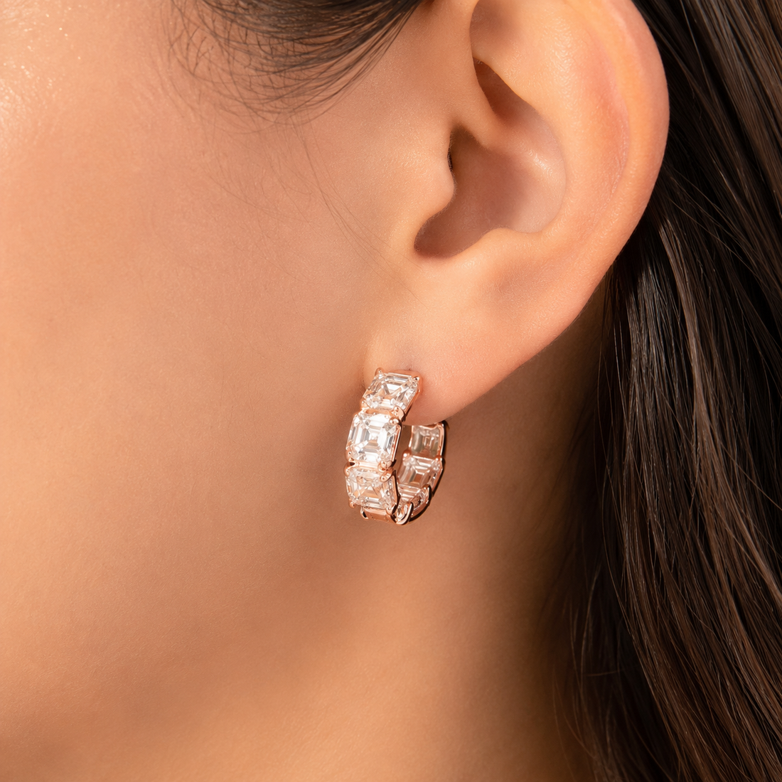 A close-up of a model's ear wearing a rose gold huggie hoop earring adorned with baguette-cut clear stones.
