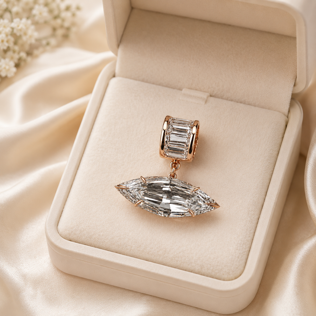 A rose gold charm with a rectangular baguette-cut clear gemstone and a dangling marquise-cut clear gemstone, displayed inside an open cream-colored jewelry box on a satin fabric background.