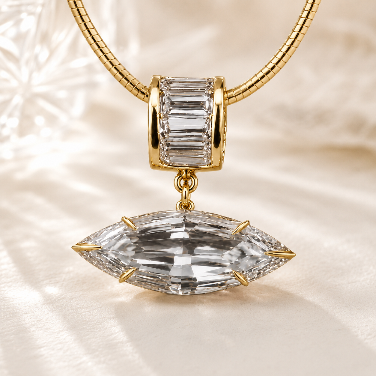 Gold-plated jewelry charm featuring a large marquise-cut clear crystal pendant and a smaller baguette-cut clear crystal accent, shown on a gold snake chain against a soft, light beige textured background.