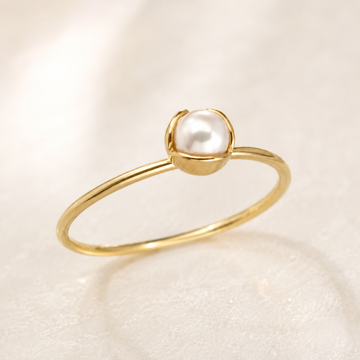 A delicate gold ring featuring a single round white pearl held in a small gold bezel setting, resting on a soft, textured white surface.