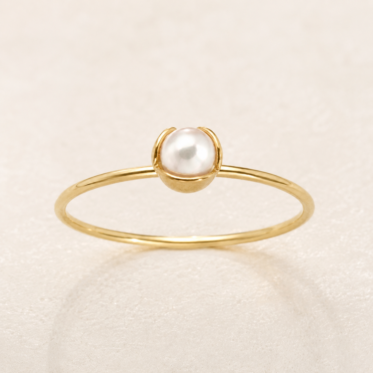 A delicate gold ring featuring a single round white pearl set in a small gold dome, photographed on a light beige textured background.
