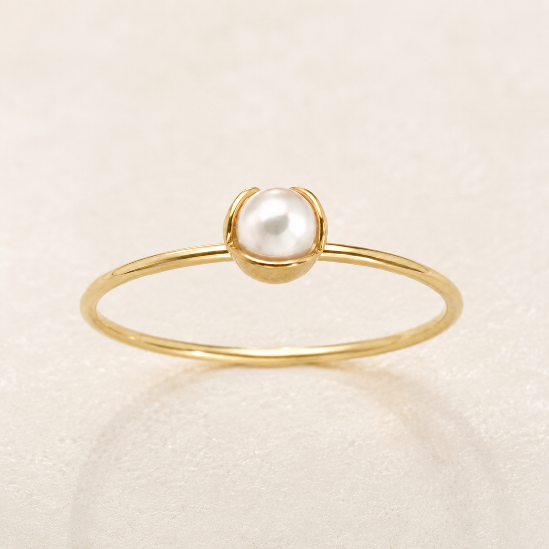 A delicate gold ring featuring a single round white pearl set in a small gold dome, photographed on a light beige textured background.