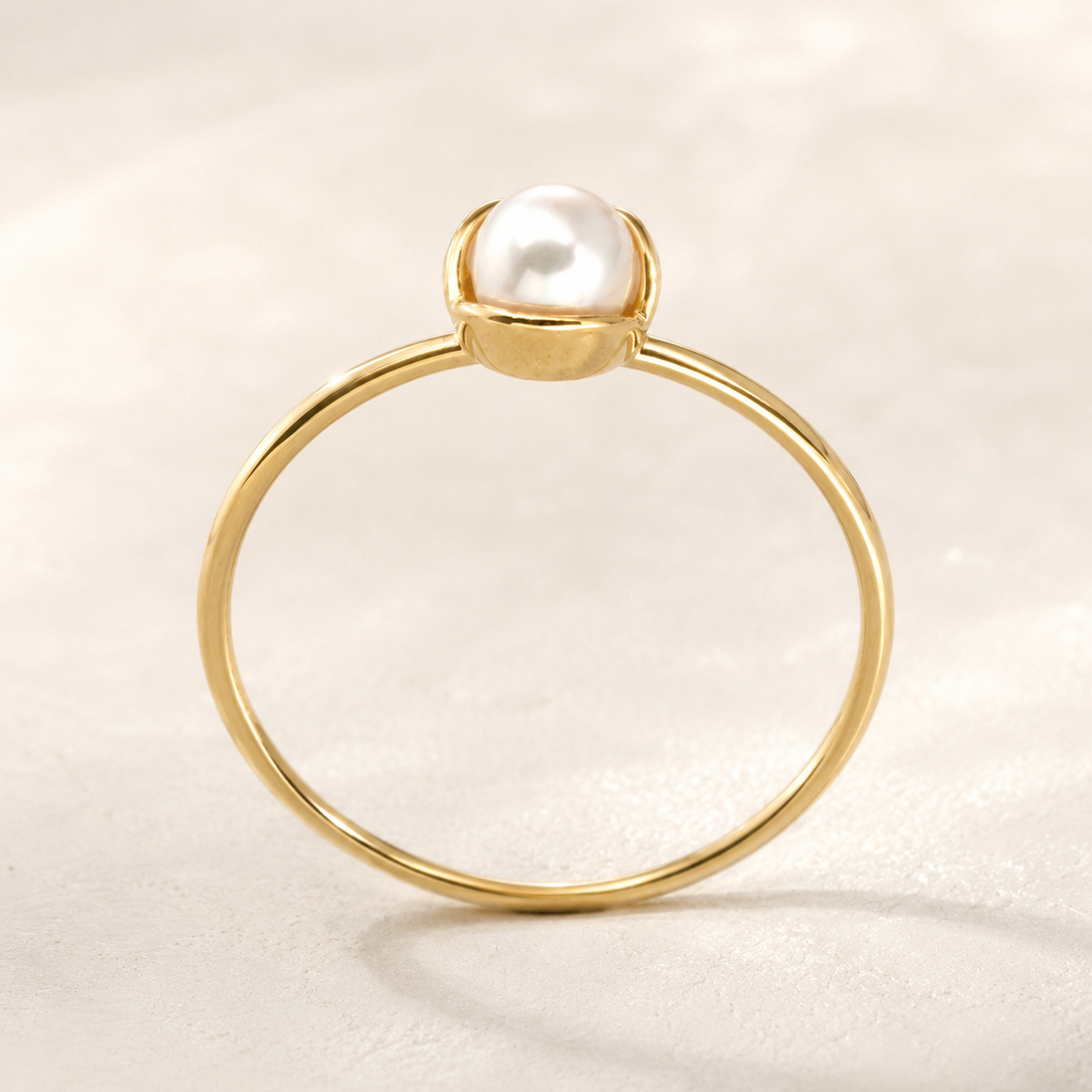 A delicate gold ring featuring a single white pearl set in a small bezel, displayed on a soft, light beige textured surface.