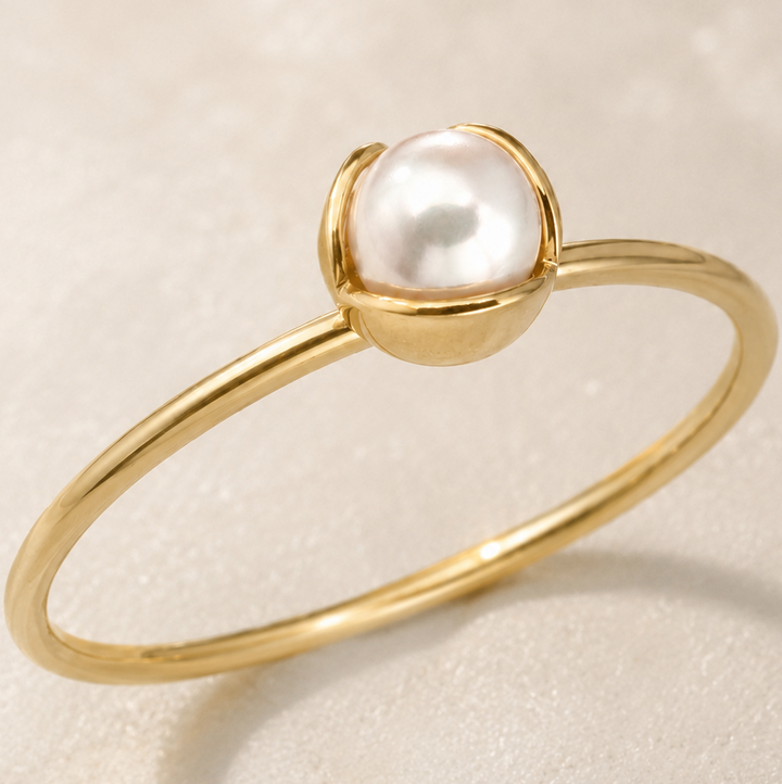 A delicate gold ring featuring a single white pearl set in a small gold dome, photographed on a soft, light beige textured background.