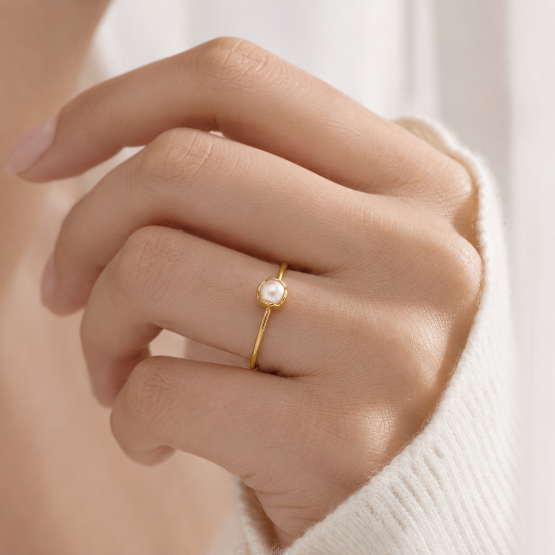 A close-up of a hand wearing a delicate gold ring with a small white pearl bezel-set on a soft, light-colored knit sweater.