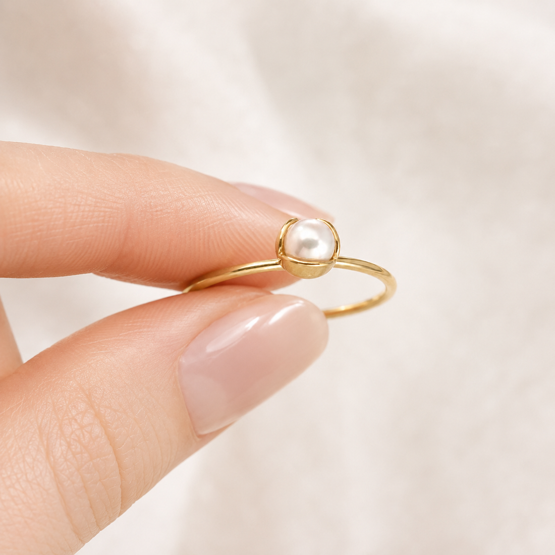 A delicate gold ring with a single white pearl bezel-set on a person's finger, photographed against a soft, light beige fabric background.