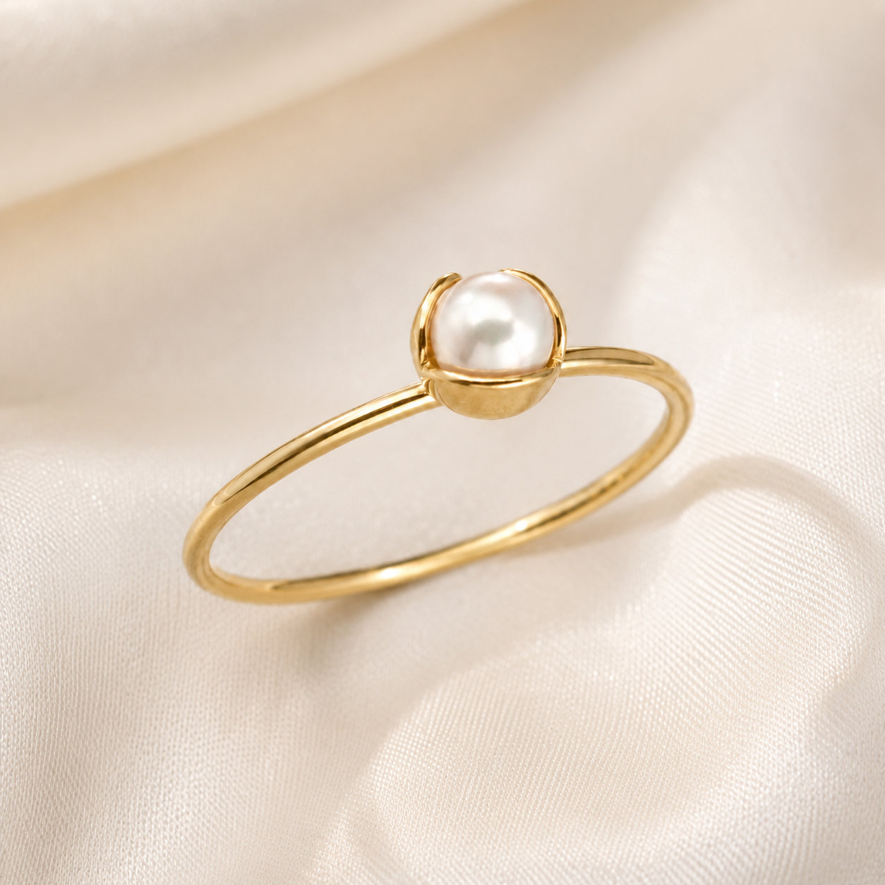 A delicate gold ring featuring a single white pearl set in a small gold bezel, resting on draped cream-colored satin fabric.