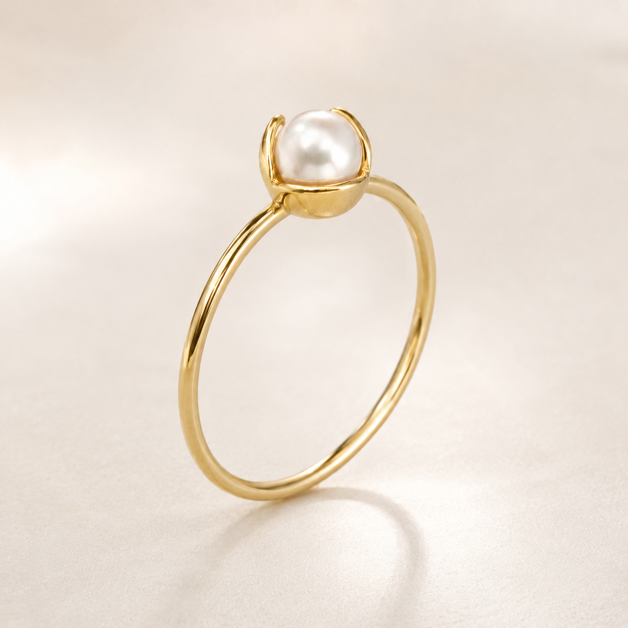 A delicate yellow gold ring featuring a single round white pearl held in a simple prong setting, photographed on a soft, light beige background with a subtle reflection.