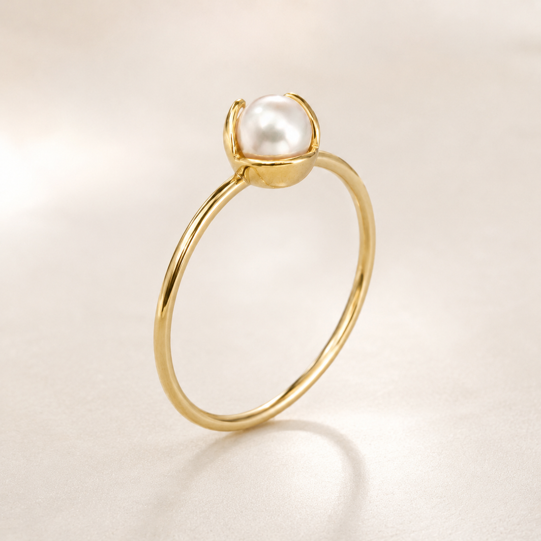 A delicate yellow gold ring featuring a single round white pearl held in a simple prong setting, photographed on a soft, light beige background with a subtle reflection.