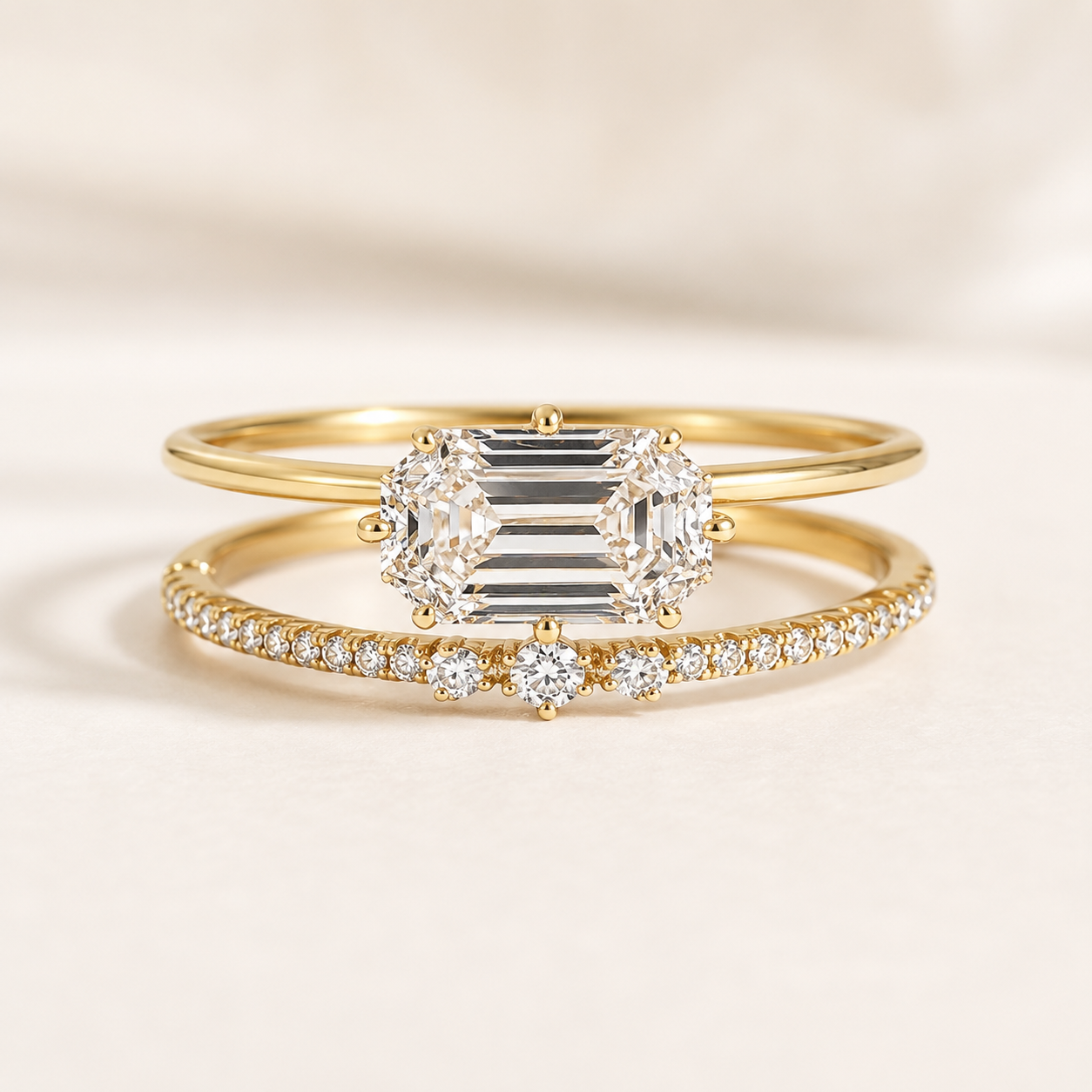 A pair of yellow gold rings, featuring a main ring with a large emerald-cut diamond and a thinner band ring paved with small round diamonds, displayed on a soft cream background.