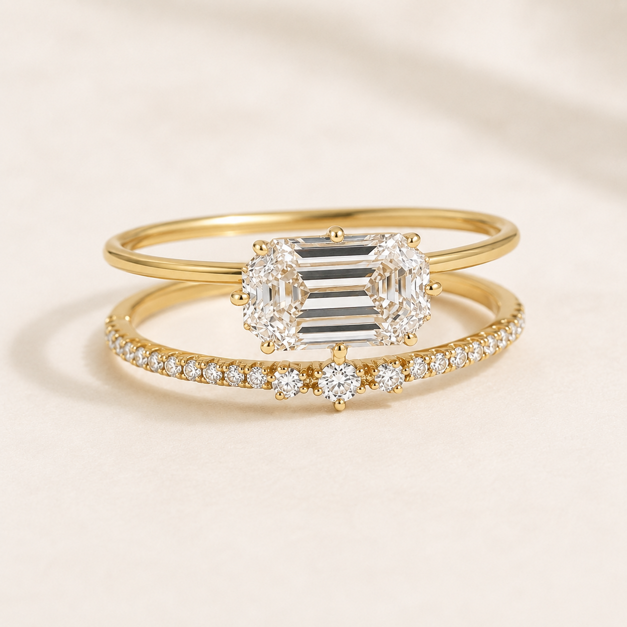 A pair of yellow gold rings displayed on a soft beige background. The top ring features a large emerald-cut diamond center flanked by round diamonds, while the bottom ring is a delicate diamond band with a small round diamond accent.