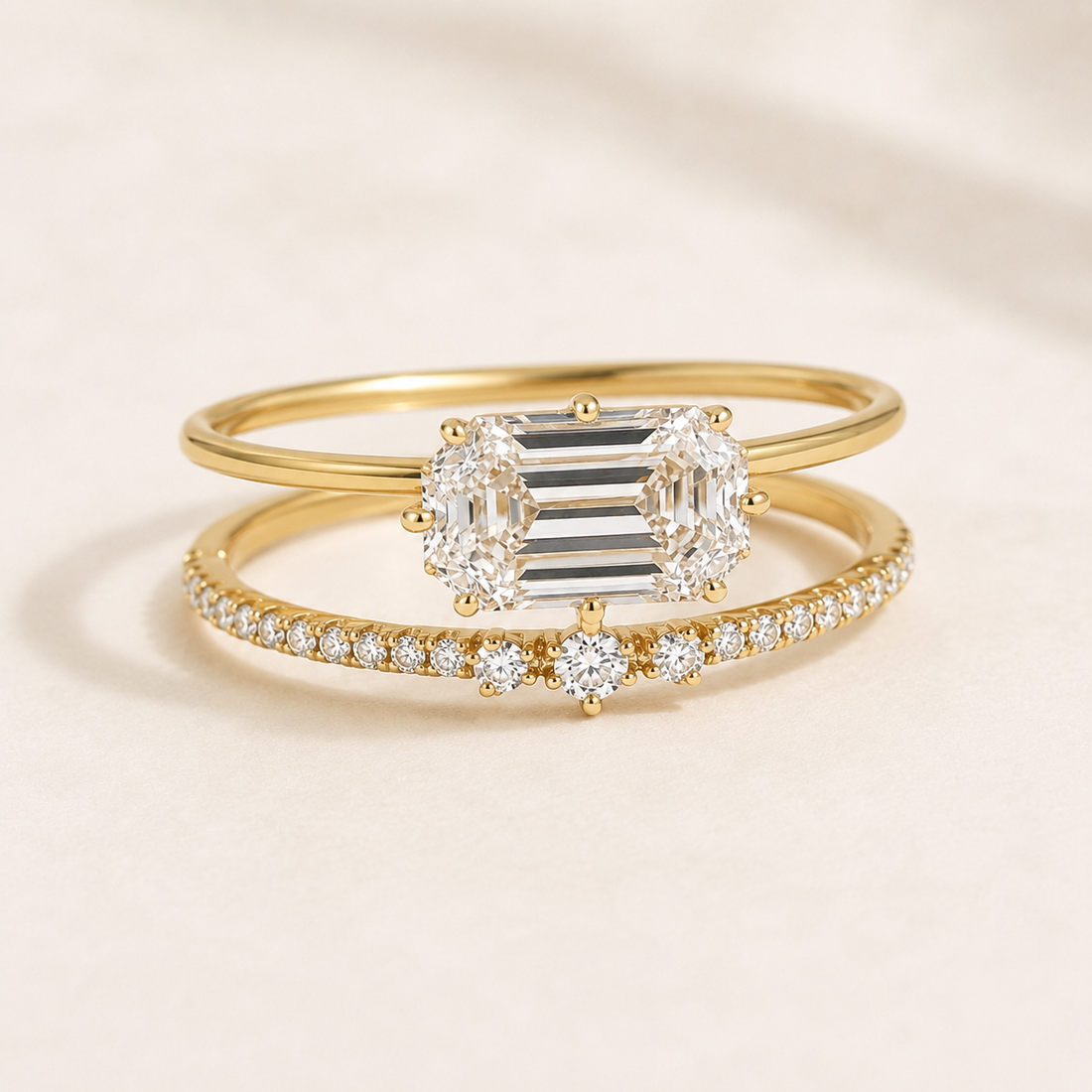 A pair of yellow gold rings displayed on a soft beige background. The top ring features a large emerald-cut diamond center flanked by round diamonds, while the bottom ring is a delicate diamond band with a small round diamond accent.