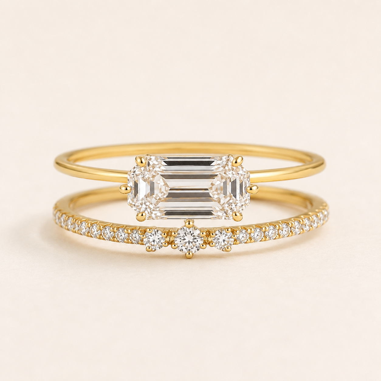 A pair of yellow gold rings with clear gemstones, displayed on a light beige background. The top ring features a large emerald-cut center stone flanked by smaller round stones, while the bottom ring is a delicate pavé band with a small round center stone.
