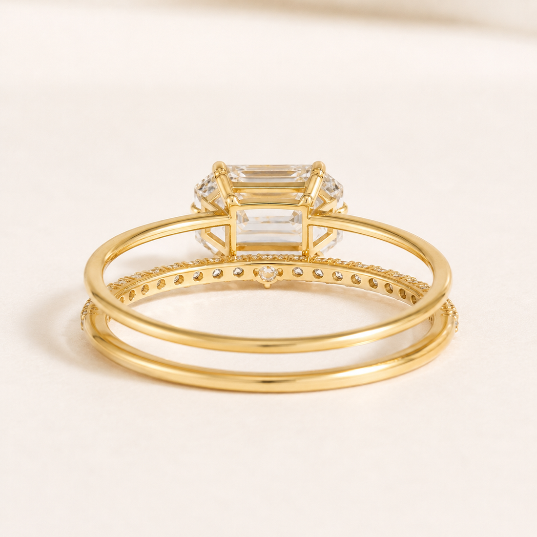 A gold double-band ring featuring a large rectangular clear emerald-cut center stone and a row of small pavé-set stones along the band, shown on a light cream background.