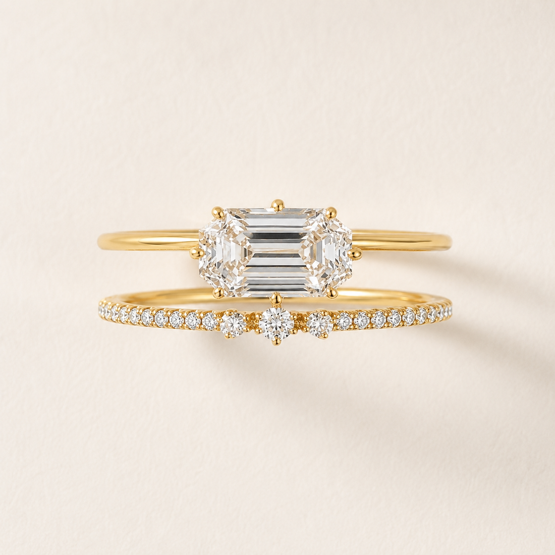 A gold ring set featuring a central emerald-cut diamond engagement ring and a matching diamond band with small pavé diamonds, displayed on a light beige background.