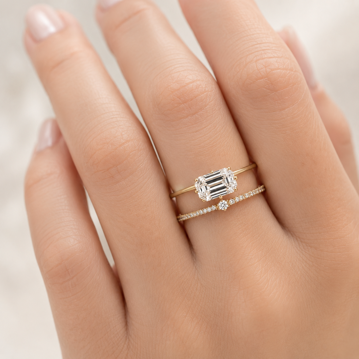 A close-up of a hand wearing a delicate gold ring set with baguette-cut clear stones and a smaller pavé band with a central round stone, photographed against a soft light background.
