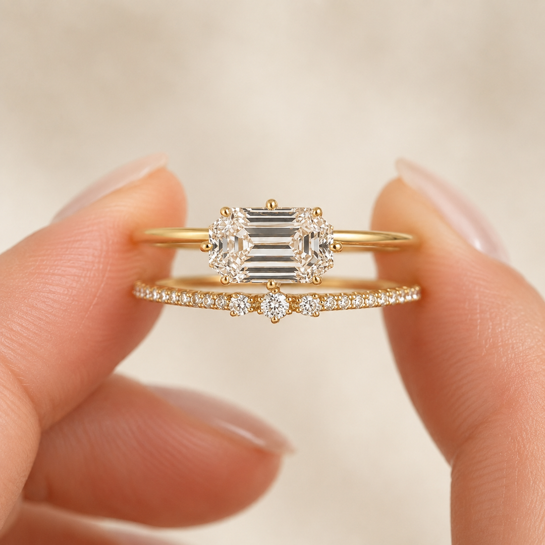 A pair of yellow gold rings held between fingers against a soft beige background. The top ring features a central baguette-cut diamond flanked by round diamonds, while the bottom ring is a delicate pavé band with a small center stone.