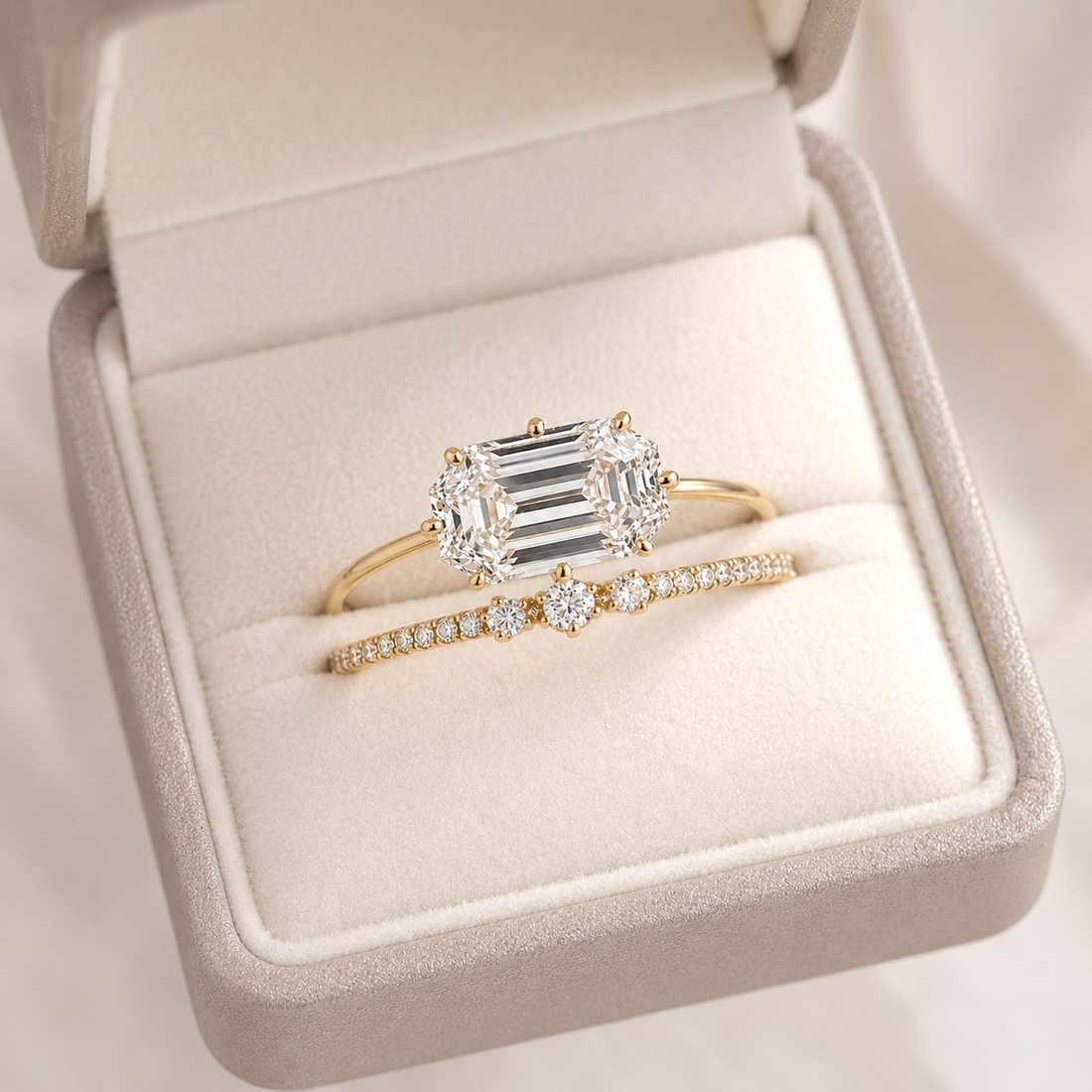 A gold ring set displayed in a beige jewelry box, featuring a yellow gold ring with a large emerald-cut center stone and two smaller round stones, alongside a matching gold band with pavé-set round stones.