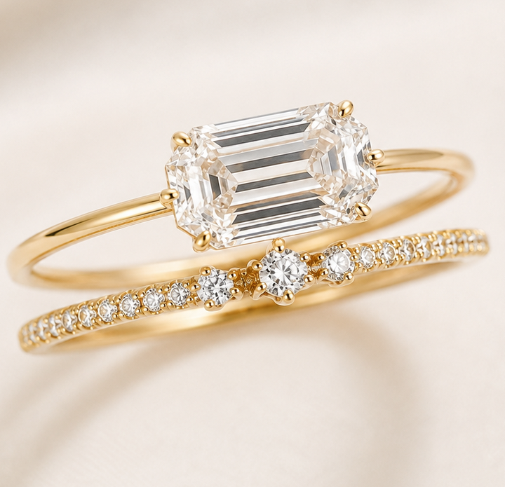 A gold ring set featuring a yellow gold engagement ring with an emerald-cut clear center stone and a matching yellow gold wedding band with pavé-set clear stones, displayed on a soft cream-colored fabric background.