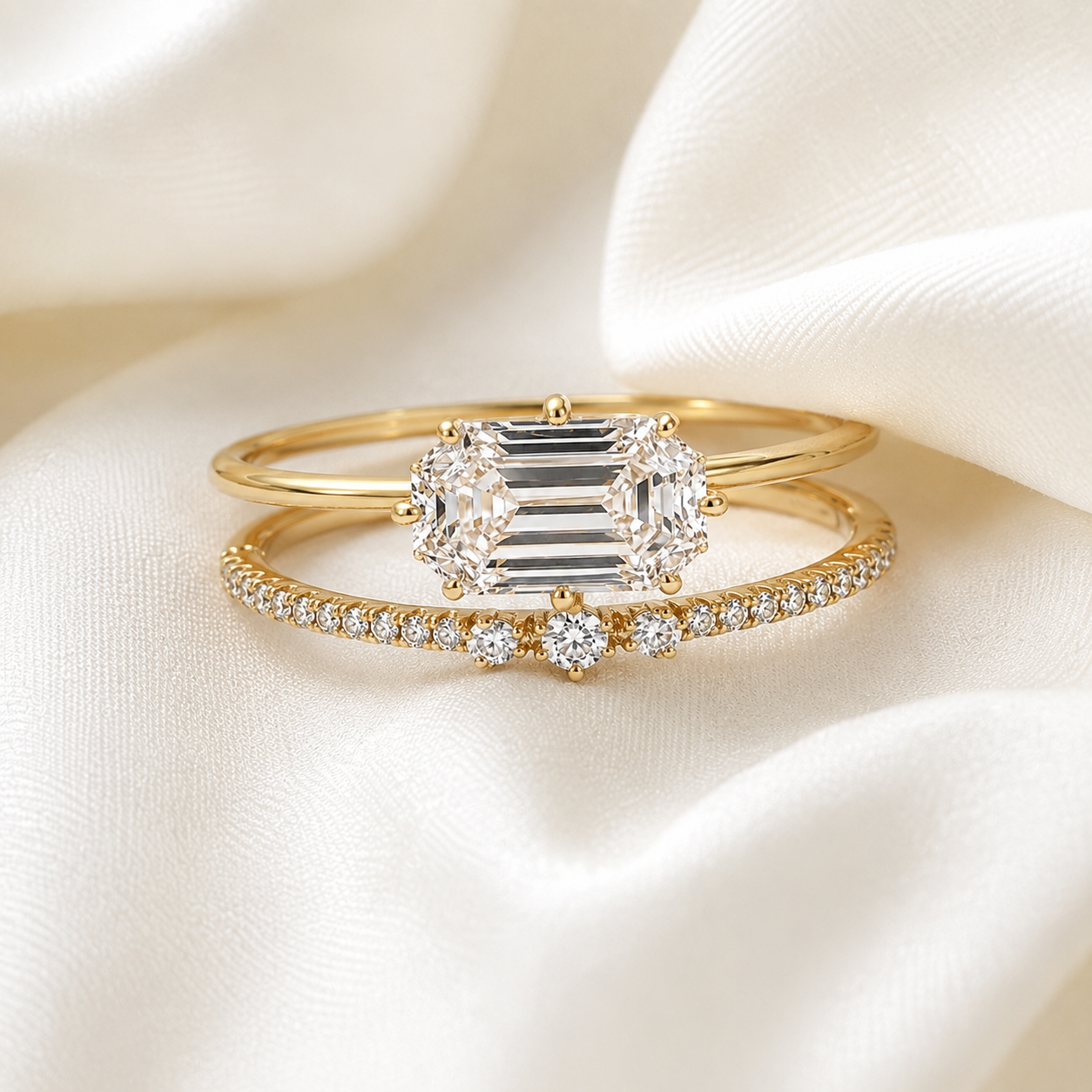 A gold ring set featuring a central emerald-cut diamond cluster flanked by two smaller diamonds, displayed on a delicate gold band paved with small round diamonds, all resting on draped white satin fabric.