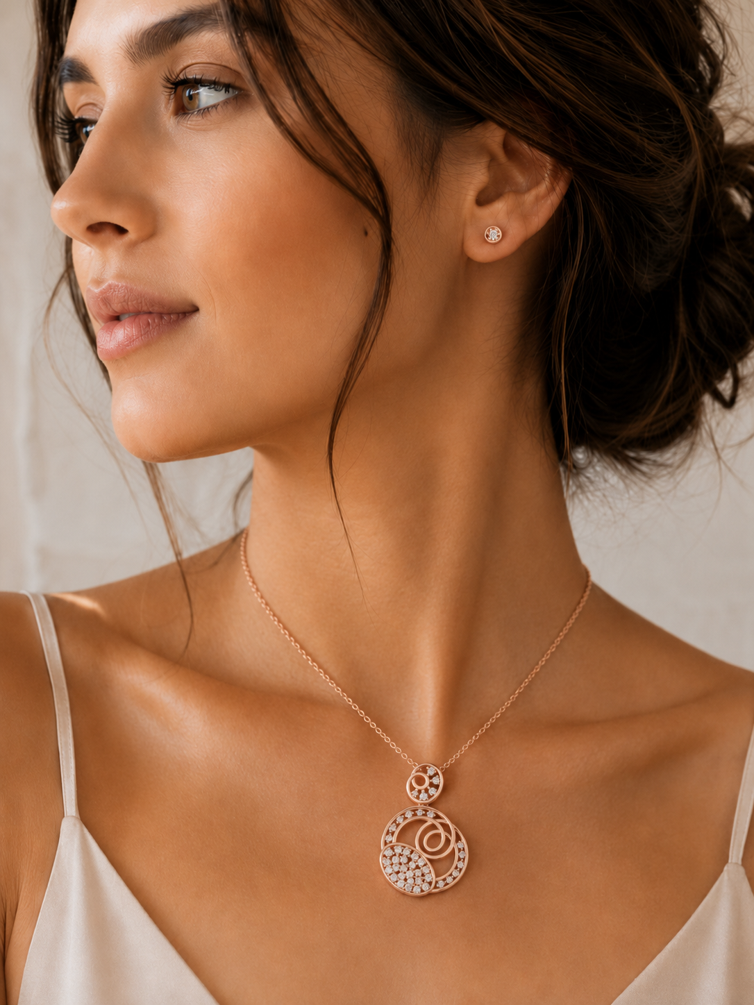 A close-up lifestyle shot of a woman wearing a rose gold circular pendant necklace with clear crystals and a matching rose gold stud earring.