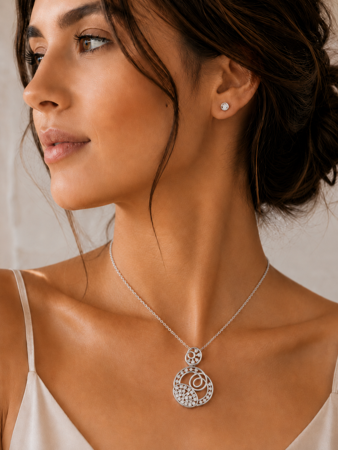 A close-up of a woman wearing a silver pendant necklace with a circular, textured design and a matching stud earring, set against a soft beige background.