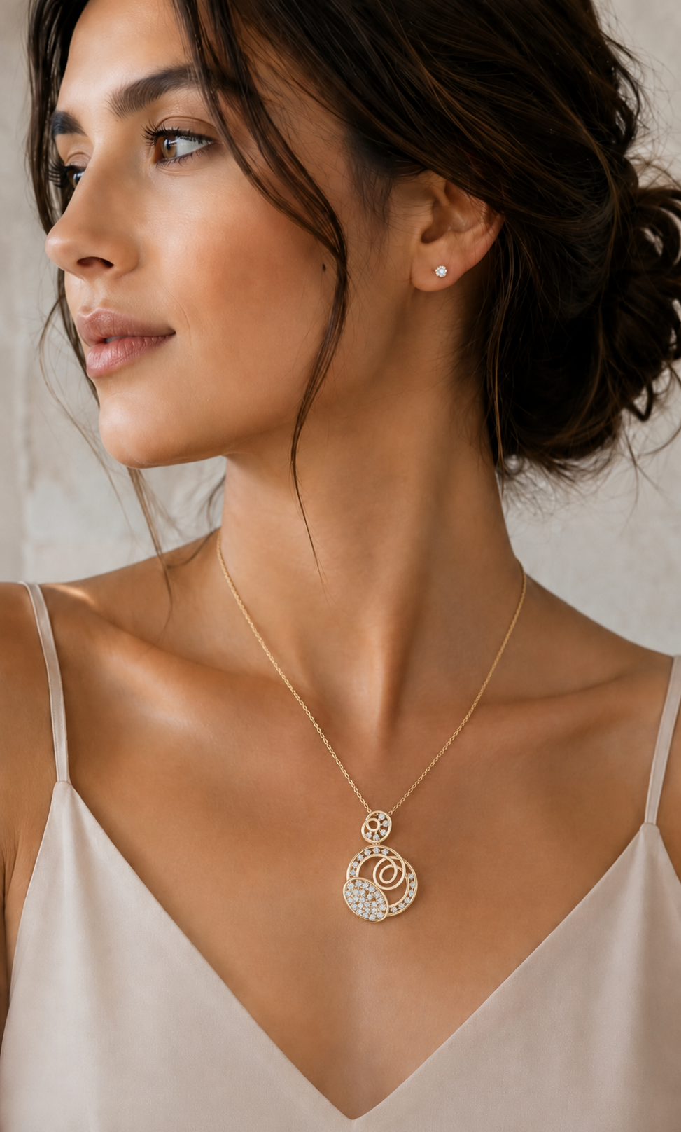A woman wearing a delicate gold pendant necklace with a circular, textured design. The necklace is shown on her neck, with her hair pulled back and a small matching stud earring visible.