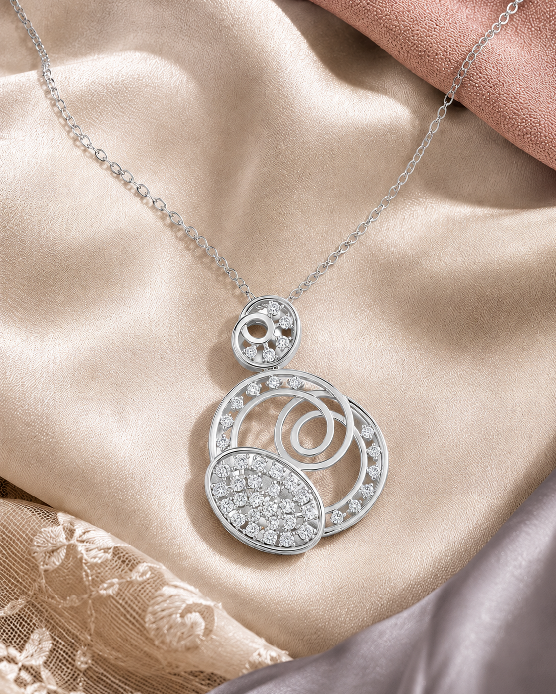 A silver-toned necklace with a multi-layered pendant featuring pavé-set clear stones, displayed on draped champagne satin fabric.