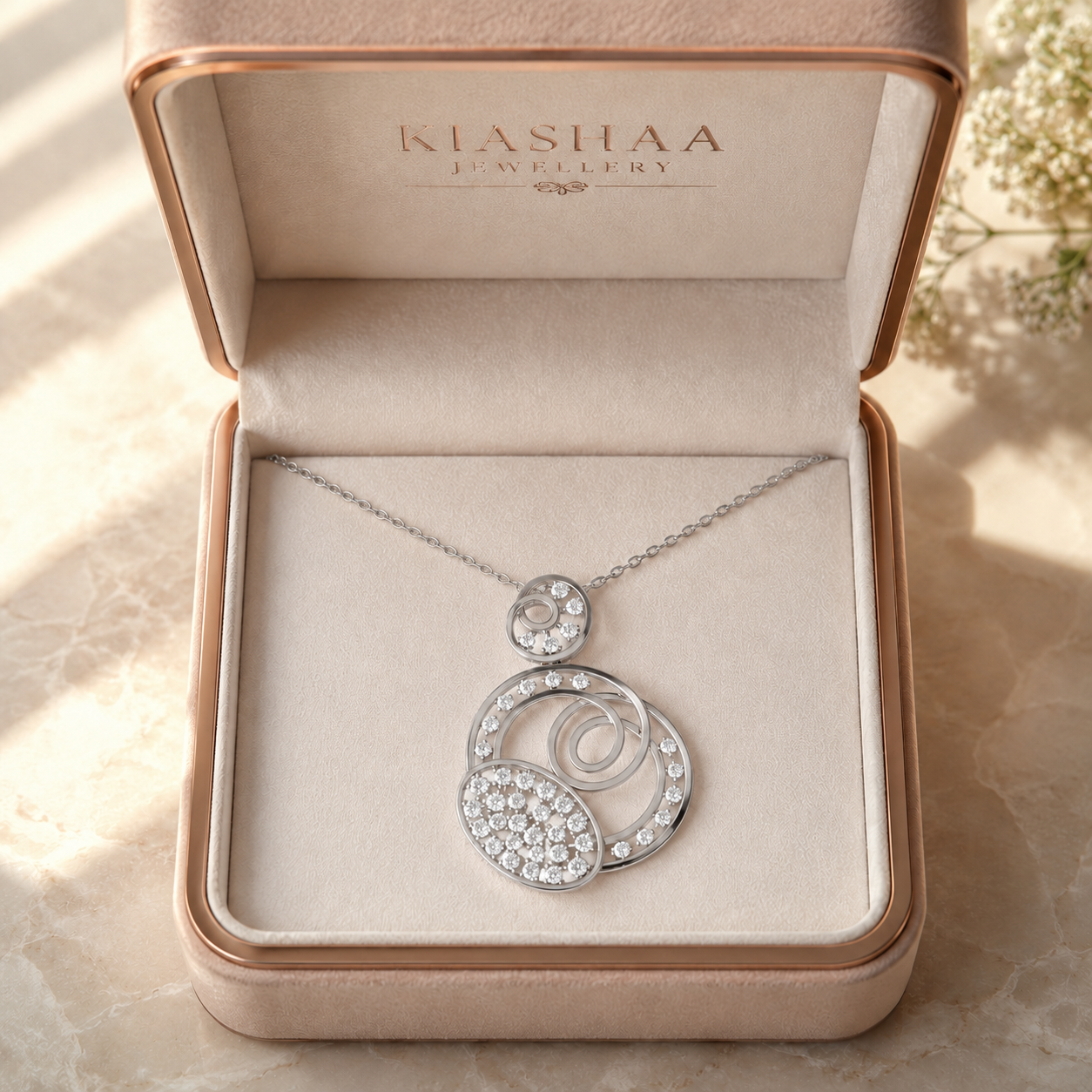 A silver pendant necklace with clear pavé and round-cut stones, displayed inside an open Klashaa Jewellery box. The necklace features a delicate chain and a multi-layered circular pendant with sparkling clear stones set in silver-toned metal, presented on a beige textured surface.