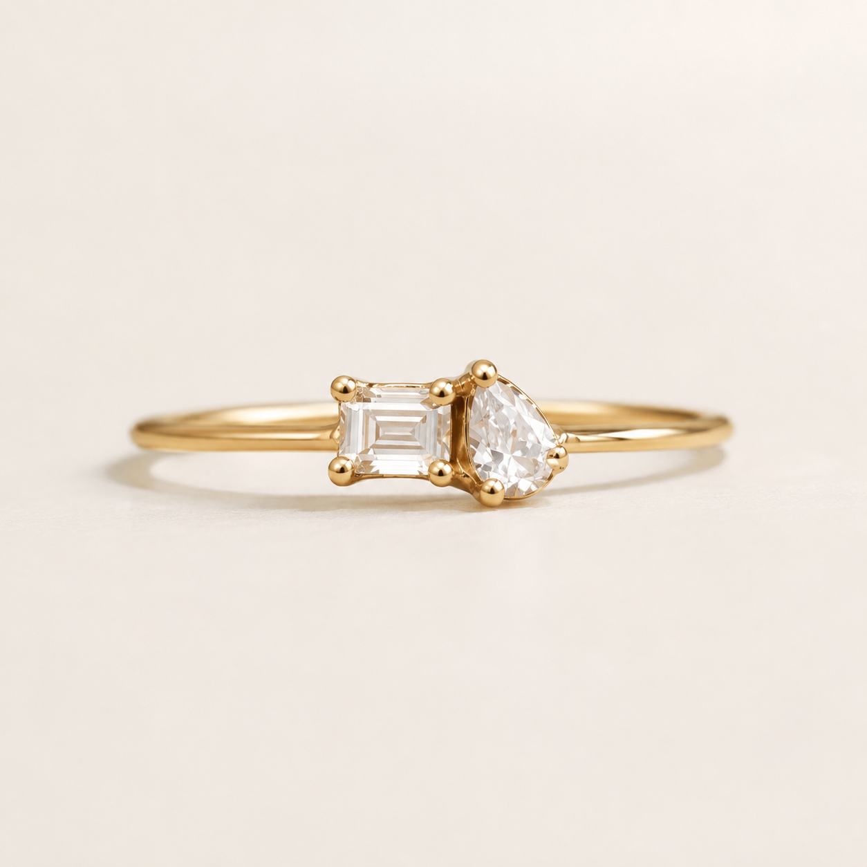A delicate gold ring featuring a pear-cut clear gemstone on one side and an emerald-cut clear gemstone on the other, set on a plain light beige background.