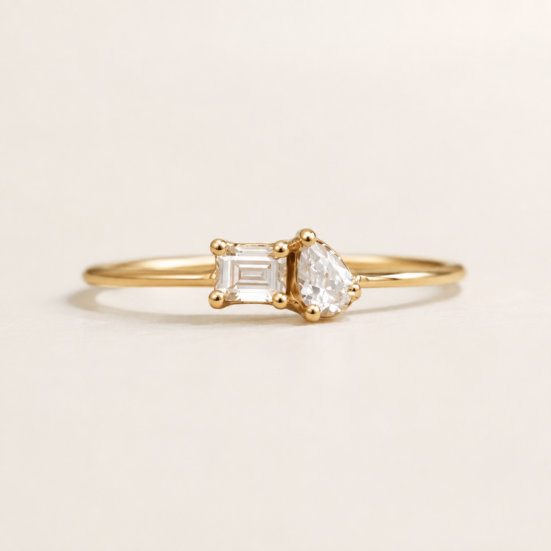 A delicate gold ring featuring a pear-cut clear gemstone on one side and an emerald-cut clear gemstone on the other, set on a plain light beige background.