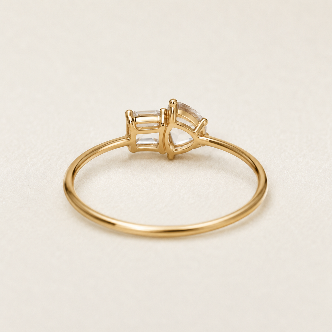 A gold ring with a bezel-set clear gemstone, photographed on a clean, light beige background.