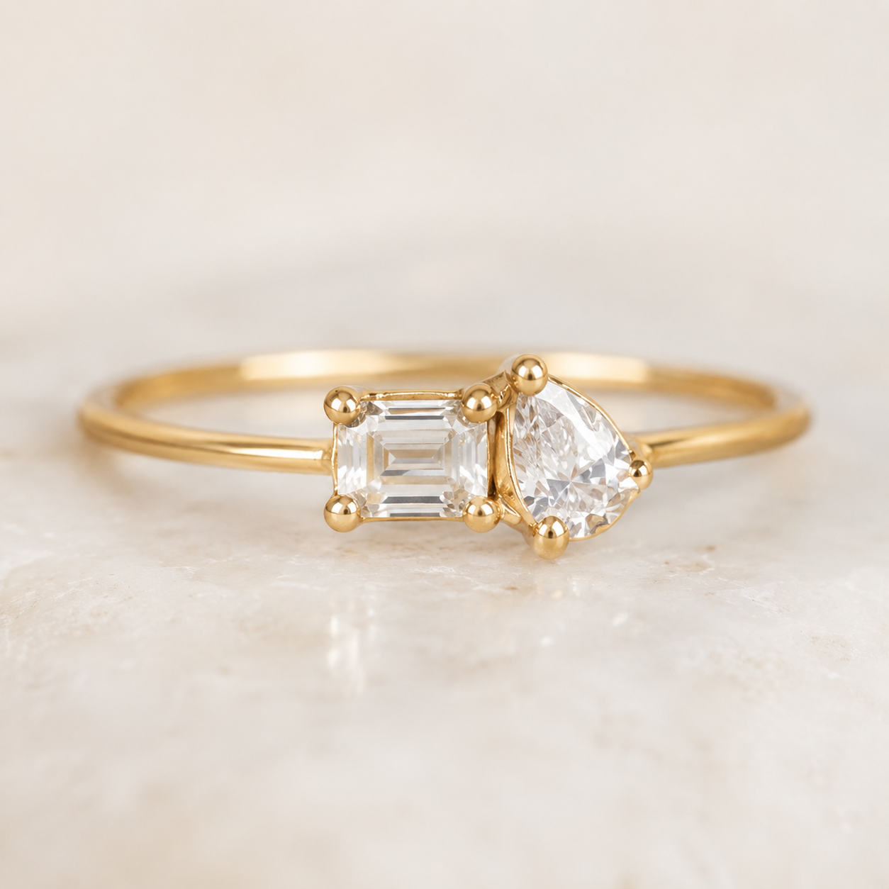 A gold ring featuring a pear-cut clear gemstone and an emerald-cut clear gemstone, displayed on a light marble surface.