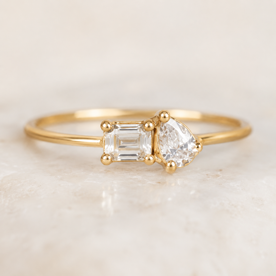 A gold ring featuring a pear-cut clear gemstone and an emerald-cut clear gemstone, displayed on a light marble surface.
