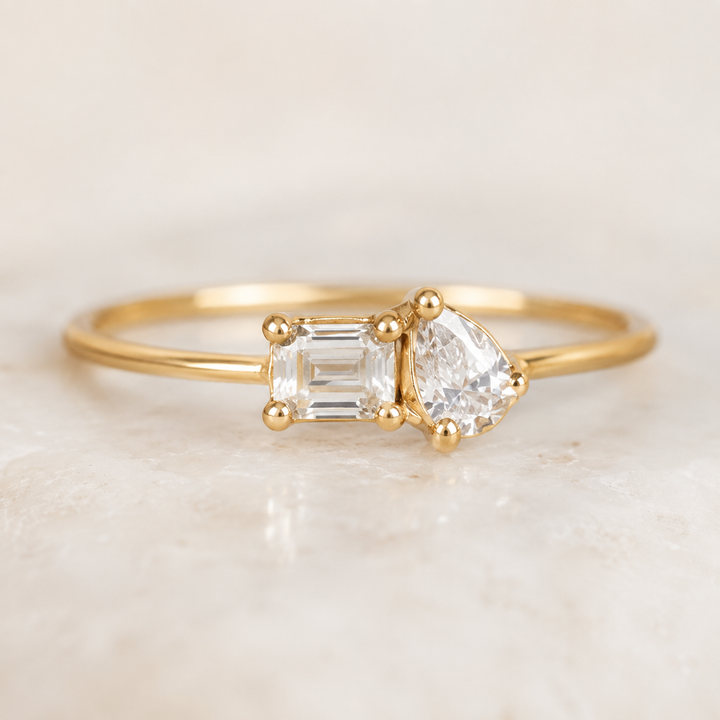 A gold ring featuring a pear-cut clear gemstone and an emerald-cut clear gemstone, displayed on a light marble surface.