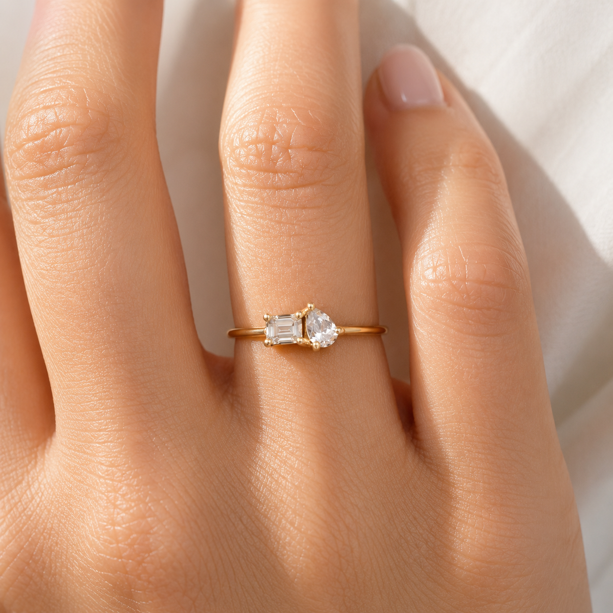 A close-up of a hand wearing a delicate gold ring with a pear-cut clear gemstone and an emerald-cut clear gemstone, set against a soft white background.