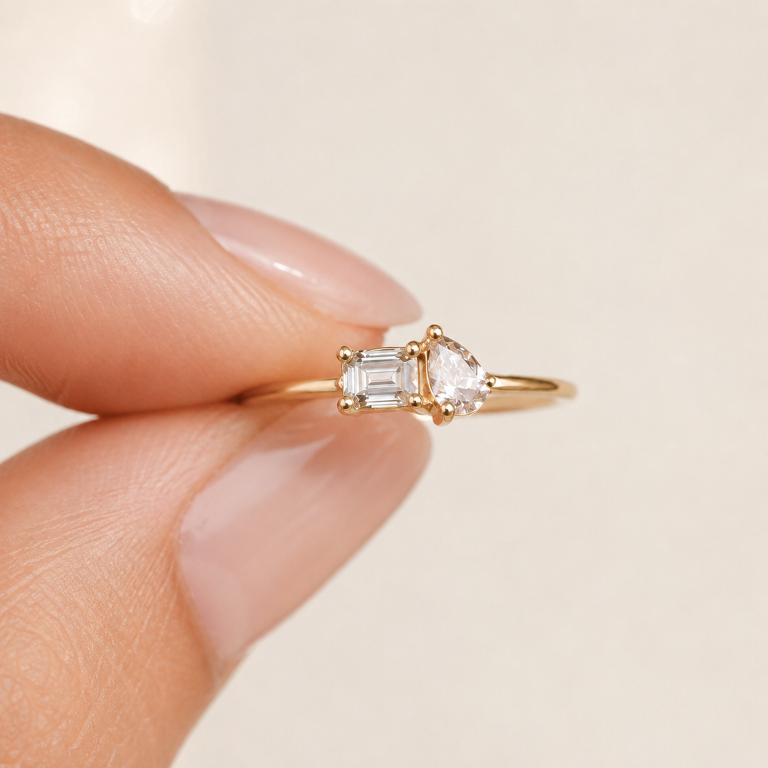 A gold ring with a pear-cut clear gemstone and an emerald-cut clear gemstone, held between two fingers against a soft beige background.