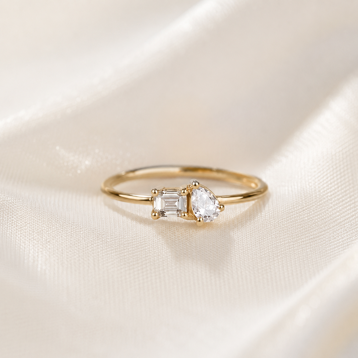 A delicate gold ring featuring a pear-cut clear gemstone and an emerald-cut clear gemstone, displayed on a soft, draped cream satin fabric.