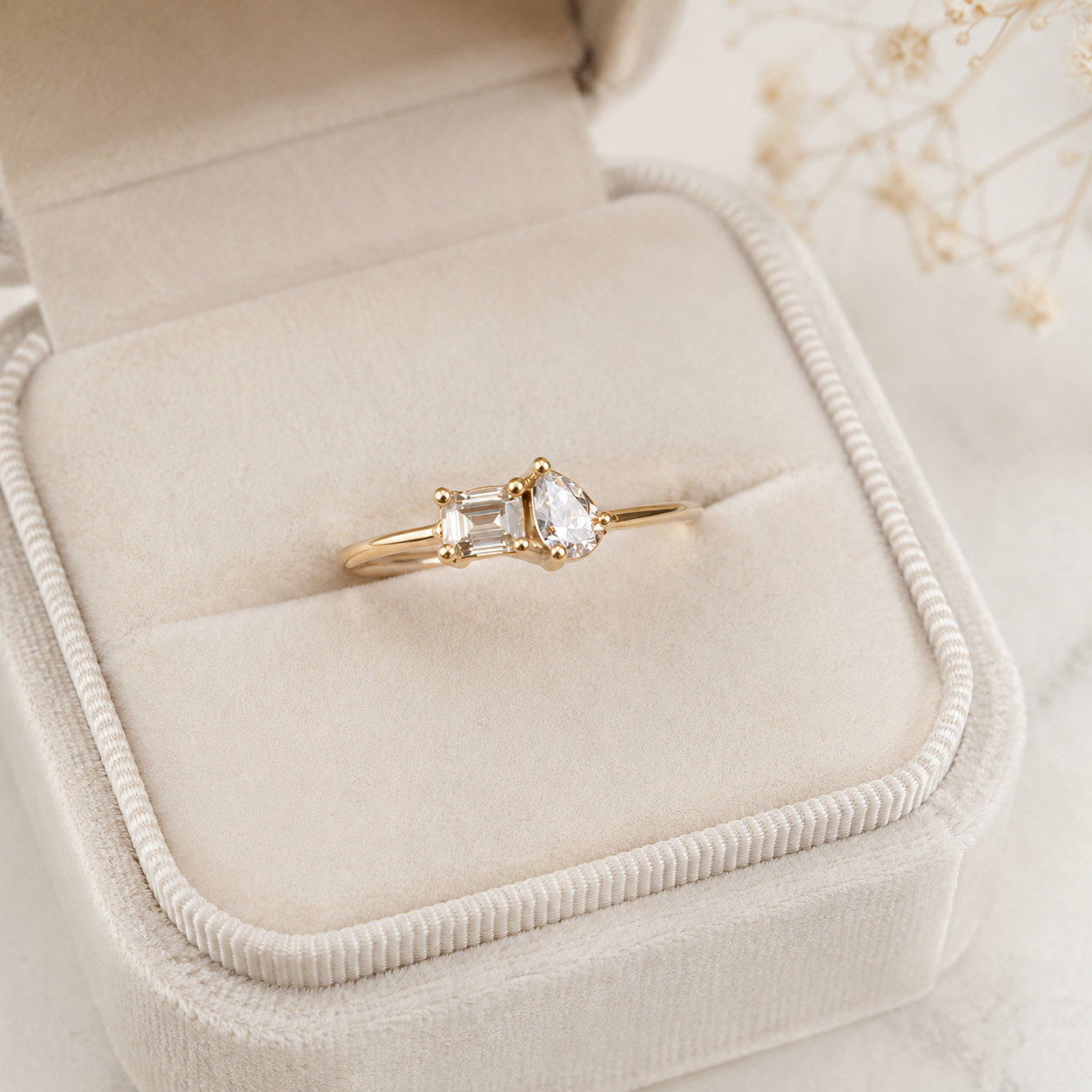 A gold ring with a pear-cut clear center stone and a rectangular clear side stone, displayed in an open cream-colored velvet ring box.