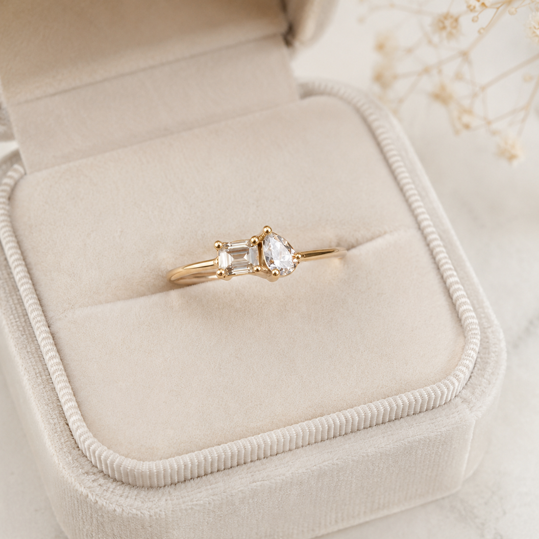 A gold ring with a pear-cut clear center stone and a rectangular clear side stone, displayed in an open cream-colored velvet ring box.