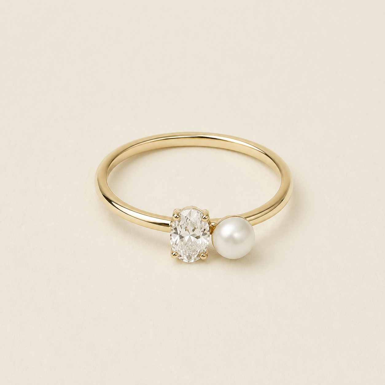 A delicate gold ring featuring a clear oval-cut gemstone and a white pearl set side by side on a thin gold band, photographed on a warm beige background.