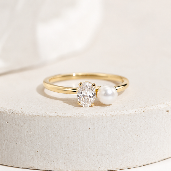 A gold ring featuring an oval-cut clear gemstone and a white pearl, displayed on a textured light gray stone pedestal against a soft white background.