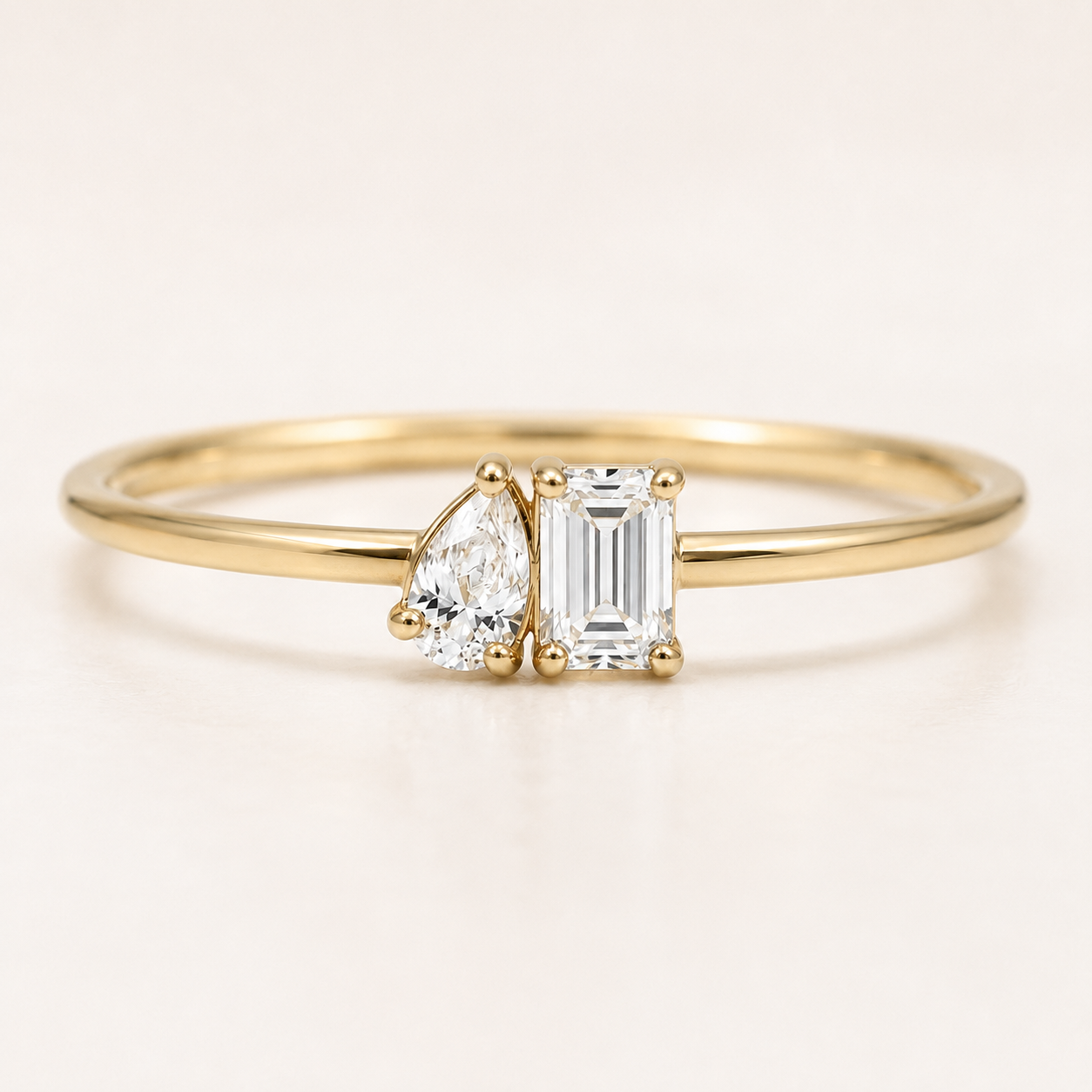 A delicate yellow gold ring featuring a pear-cut diamond and an emerald-cut diamond in a side-by-side setting on a plain light background.