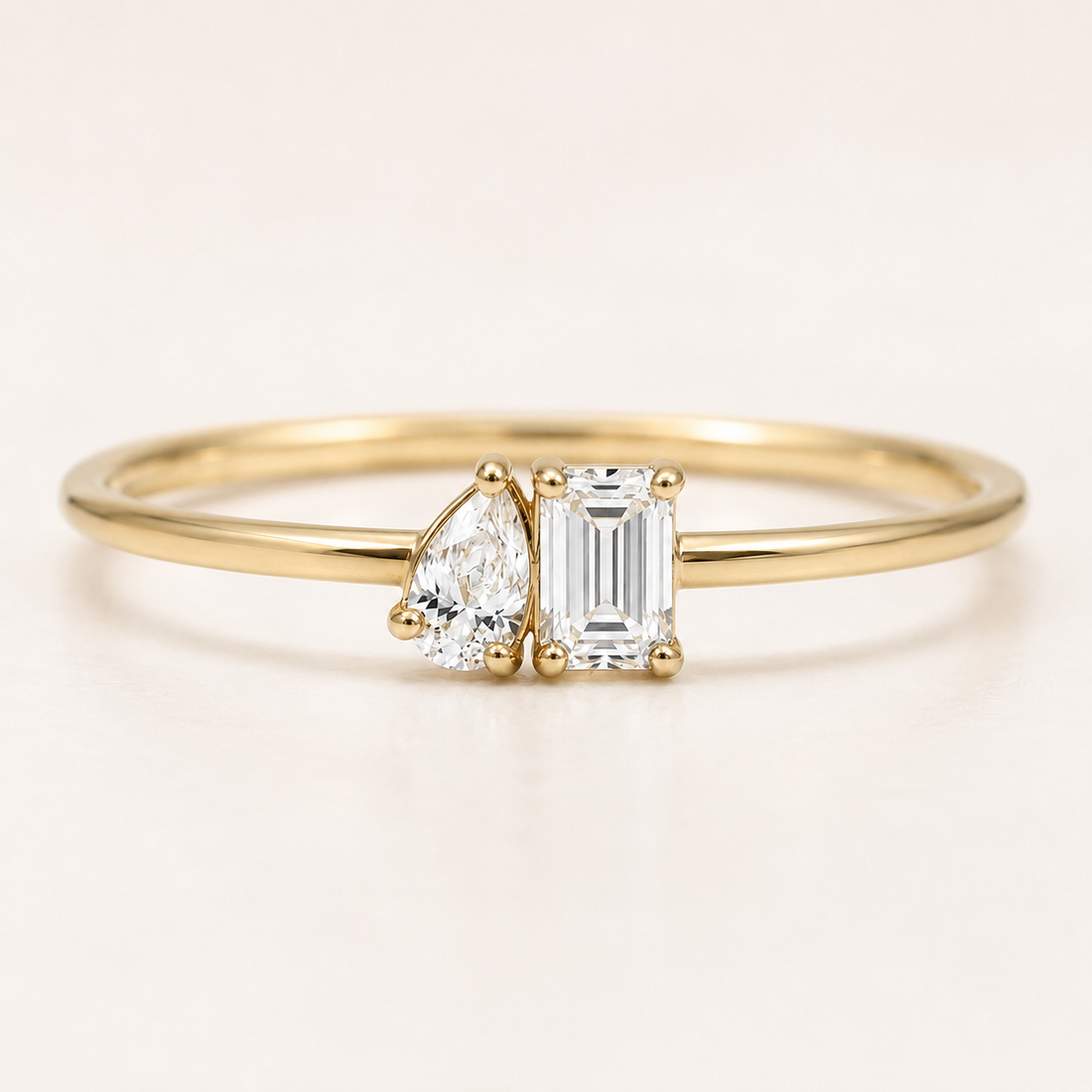 A delicate yellow gold ring featuring a pear-cut diamond and an emerald-cut diamond in a side-by-side setting on a plain light background.