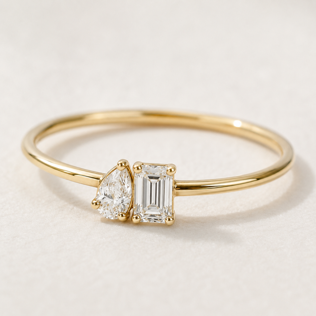 A gold ring featuring a pear-cut diamond and an emerald-cut diamond set on a polished gold band, photographed on a light textured background.