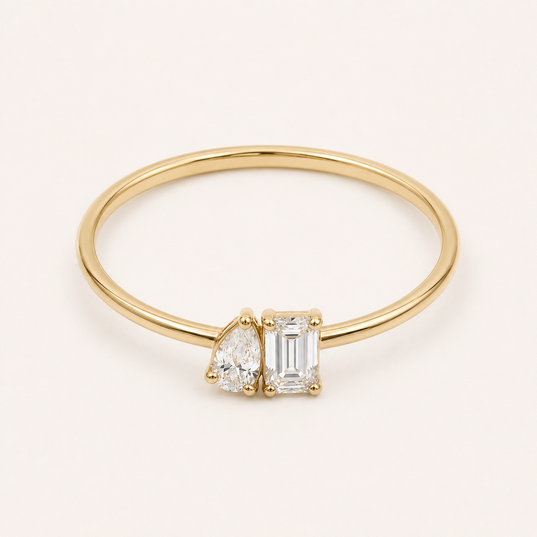 A gold ring featuring a pear-cut diamond and an emerald-cut diamond set in a polished yellow gold band, shown on a clean off-white background.