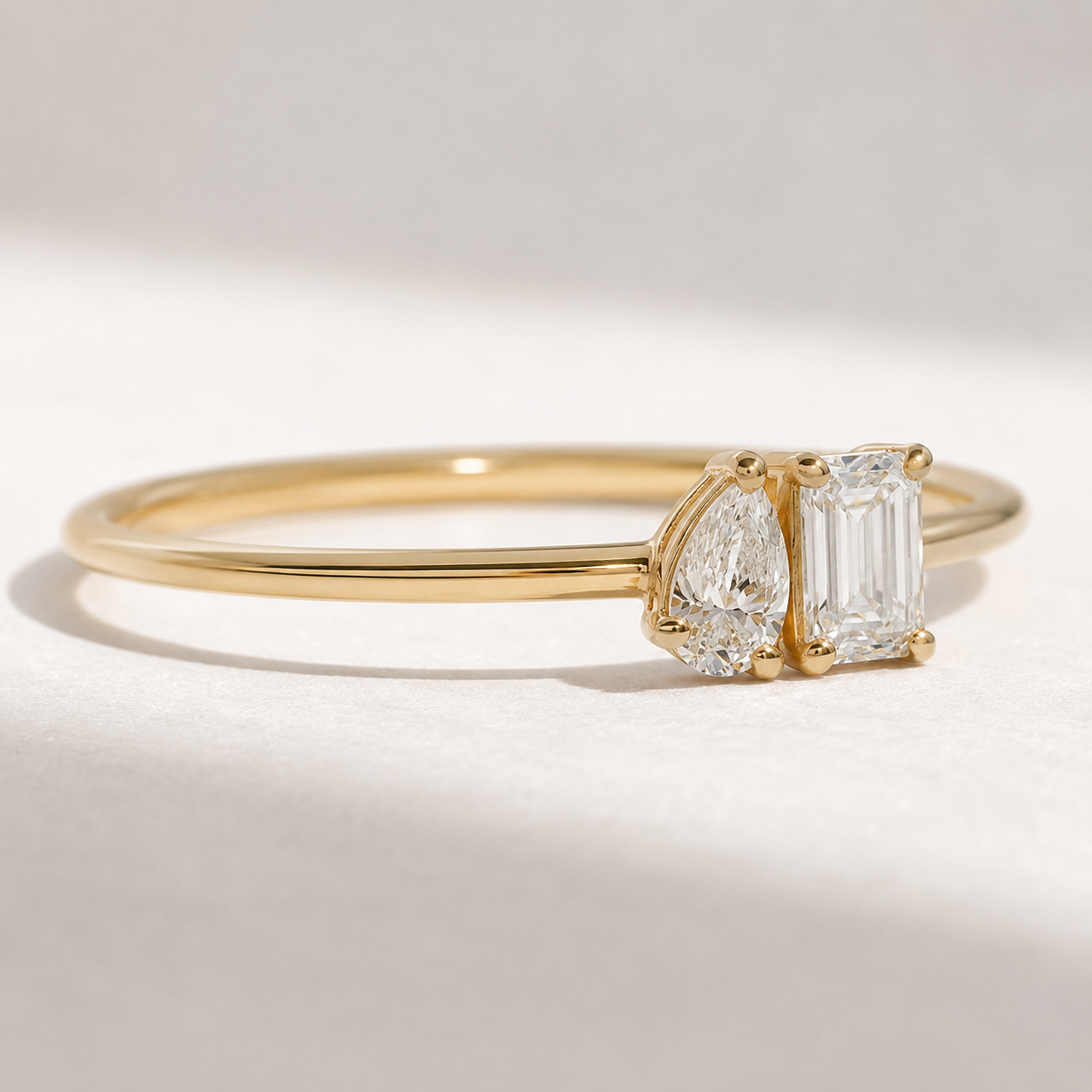 A yellow gold ring featuring a pear-cut diamond and an emerald-cut diamond, photographed on a clean, light-colored surface with soft shadows.