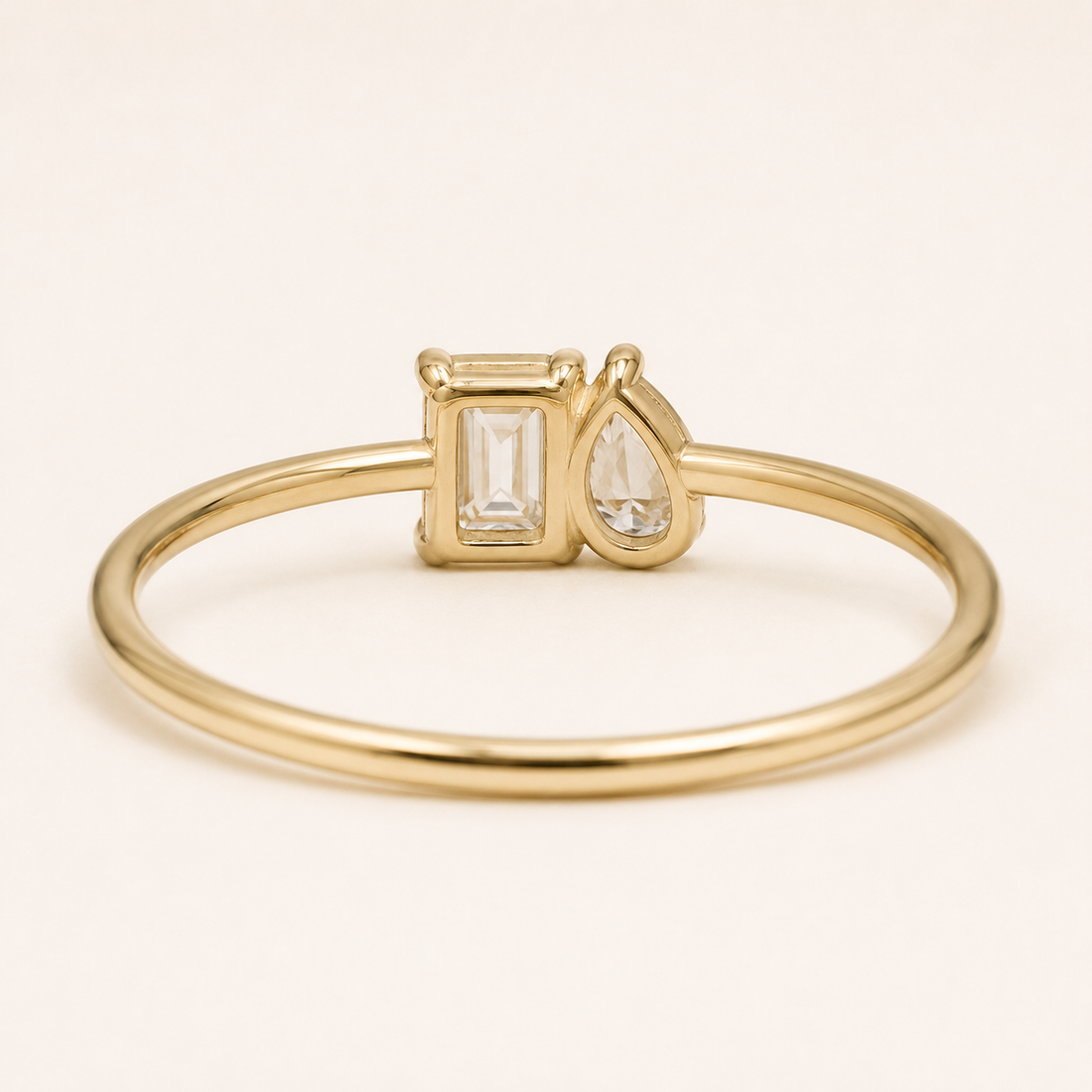 A gold ring with a slim band and a bezel-set setting featuring a rectangular baguette-cut clear gemstone and a teardrop-shaped clear gemstone, shown on a light cream background.