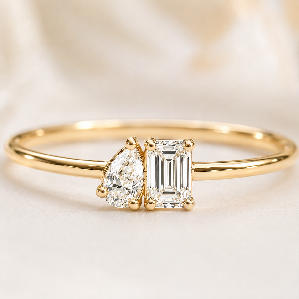 A gold ring featuring a pear-cut diamond and an emerald-cut diamond set in a four-prong setting on a polished band, photographed against a soft cream satin background.
