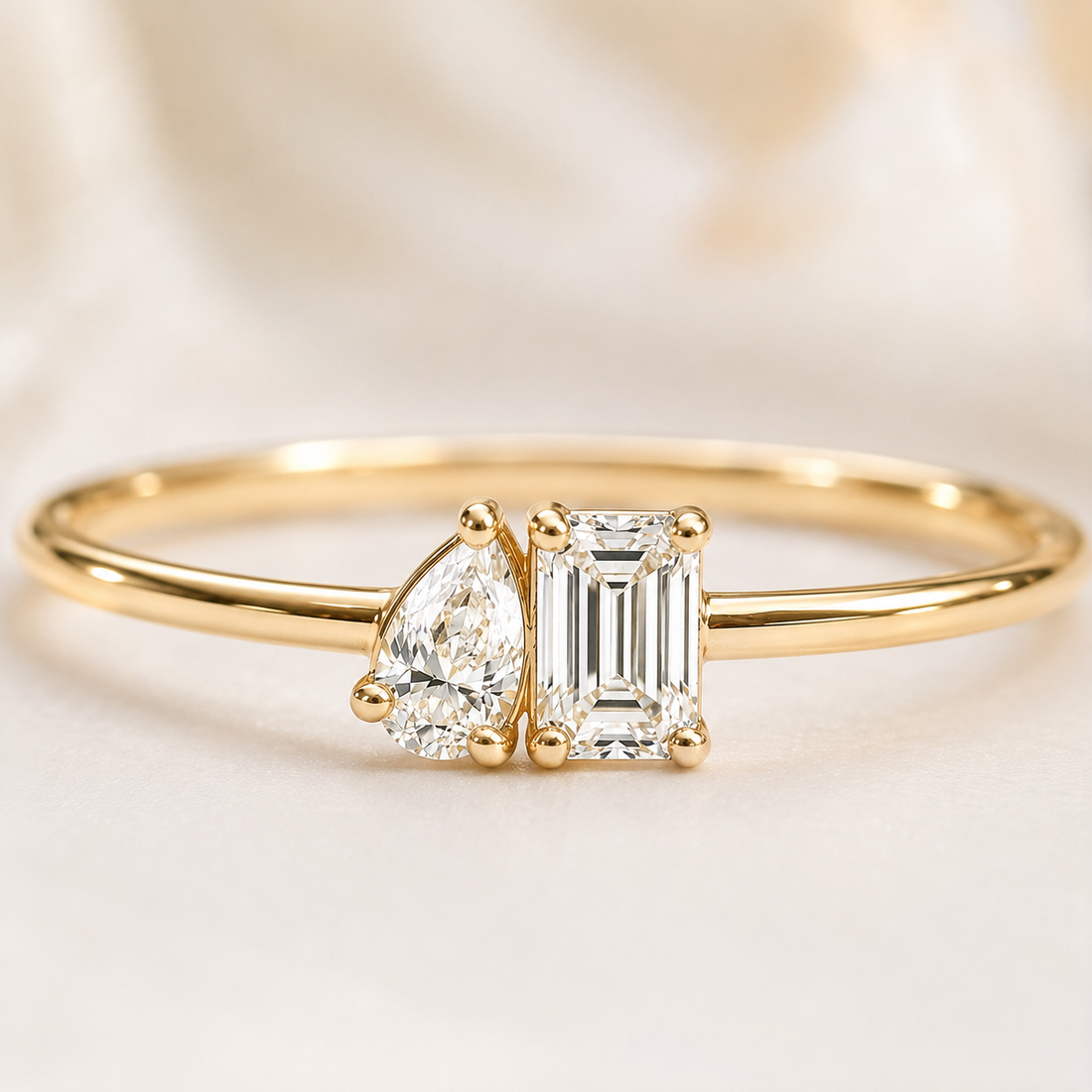 A gold ring featuring a pear-cut diamond and an emerald-cut diamond set in a four-prong setting on a polished band, photographed against a soft cream satin background.